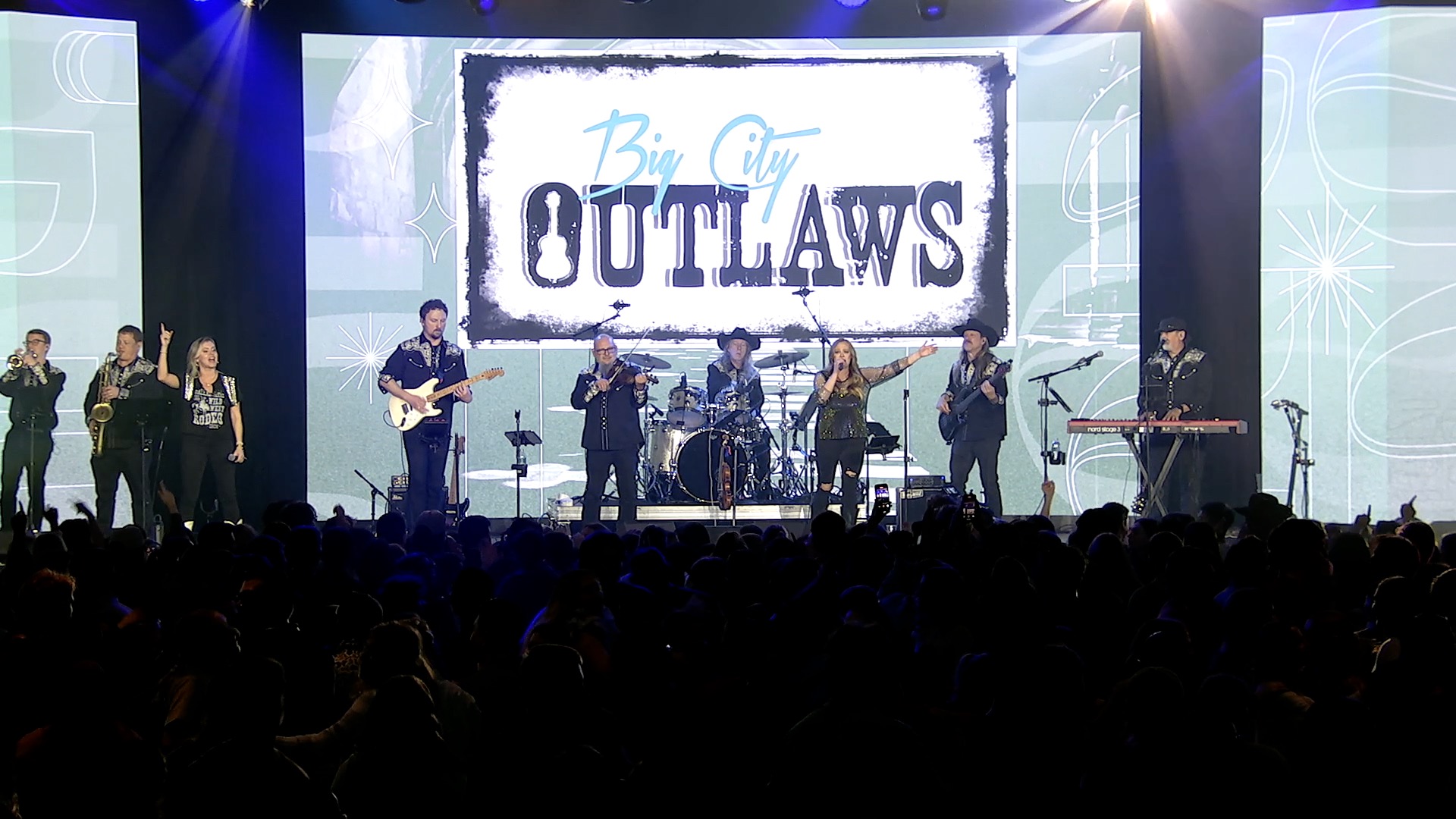 A band named "Big City Outlaws" performs on stage with guitars, brass instruments, drums, and keyboard. Bright lights shine down as a large audience watches. The Home Page Template displays the band’s name on a screen behind them.
