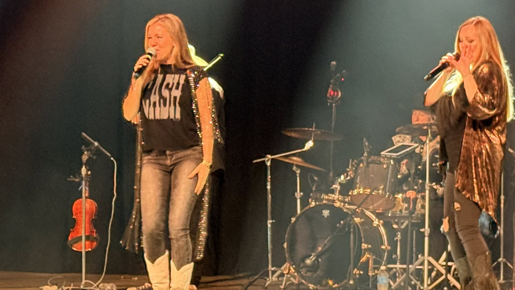 Two female singers from Dallas-Ft Worth’s #1 Country Band perform on stage with microphones, standing before a drum set and instruments, including a violin on a stand. Stage lights shine down, creating a lively concert atmosphere.