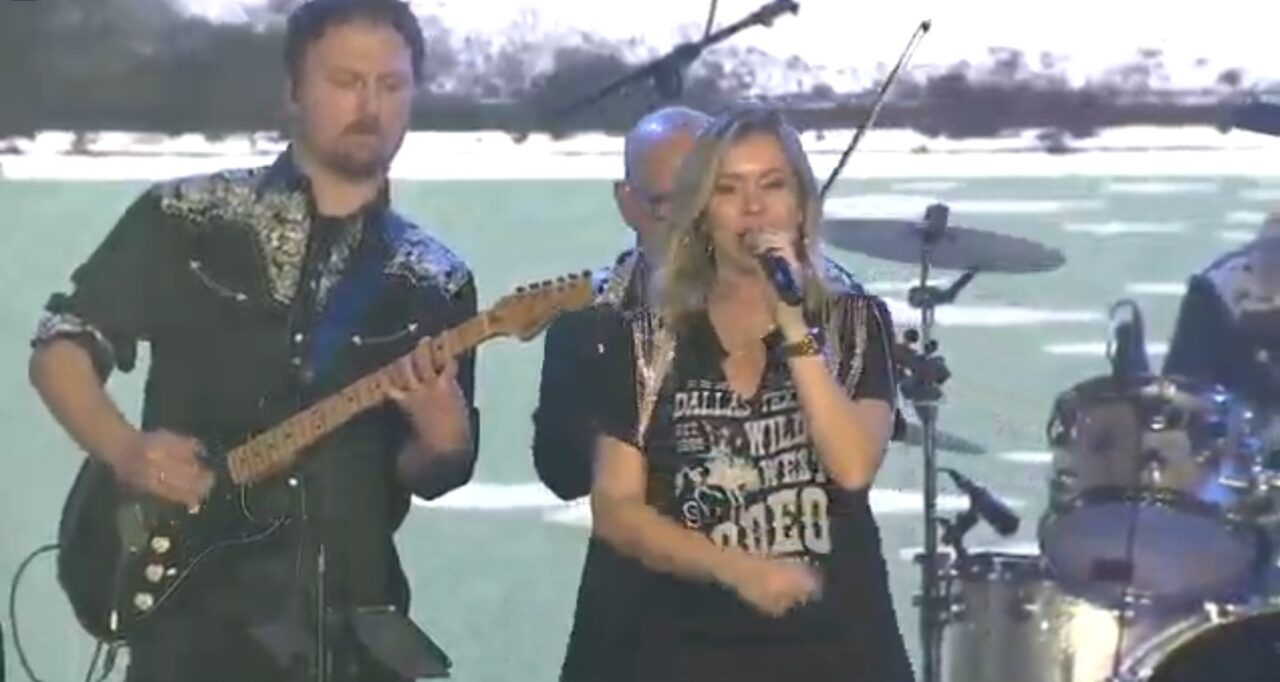 A woman sings into a microphone while a man plays electric guitar beside her on stage, with a drummer and other band equipment visible in the background.